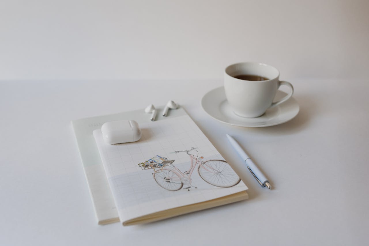 The Art of Drawing Readers In: Your attractive post title goes here A minimalist setup with a cup of coffee, notepad, pen, and wireless earbuds on a white background.